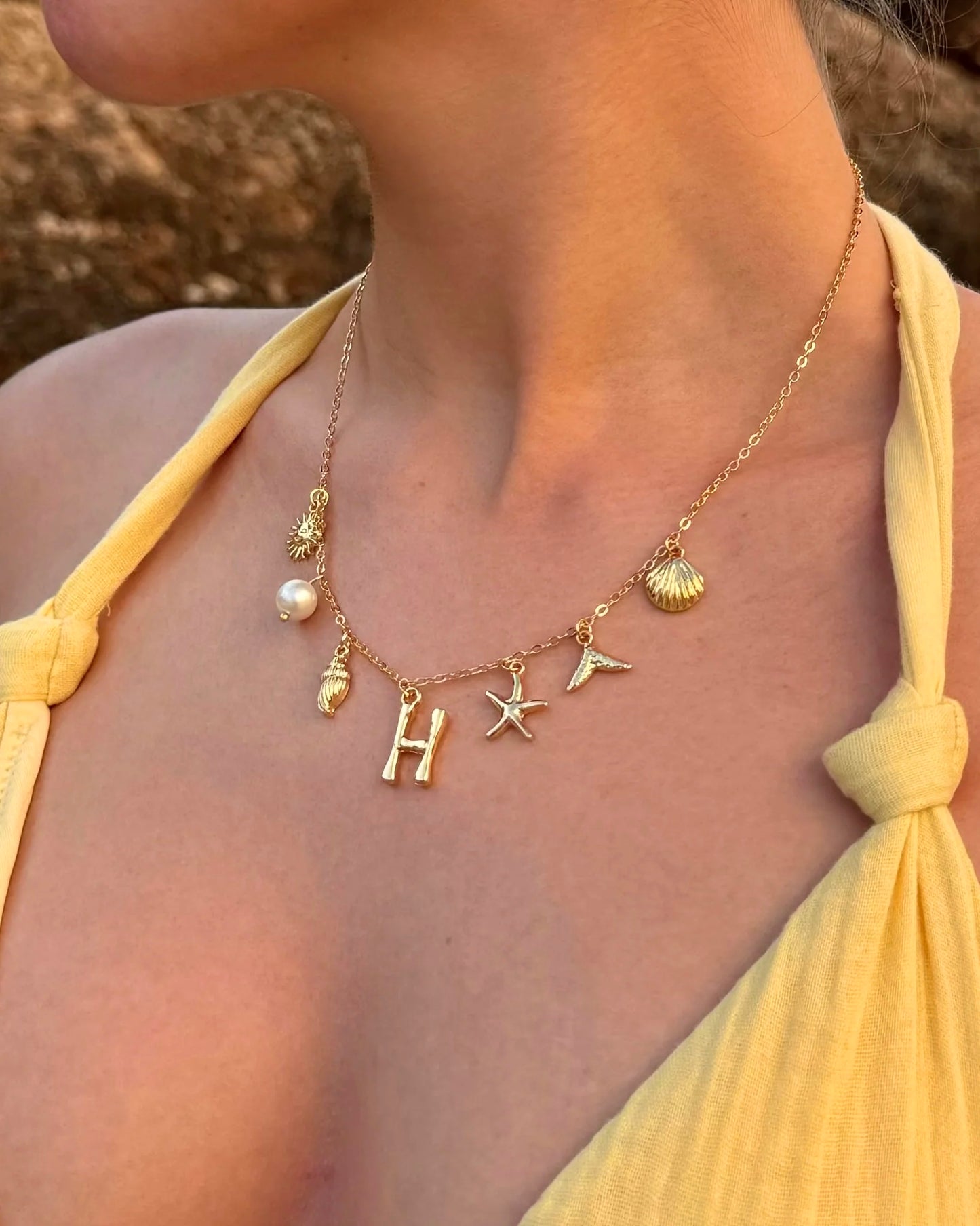Gold necklace with charms including a 'H' and sea-themed elements worn by a person in a yellow top.