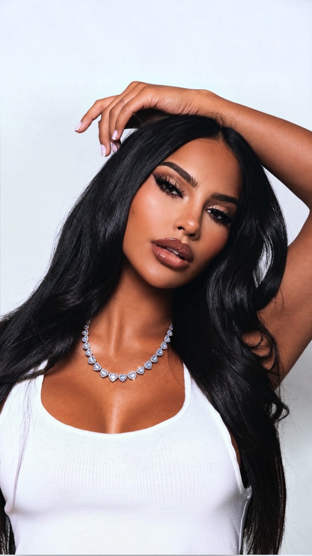 A woman with long, wavy black hair models a silver rhinestone necklace and a white ribbed tank top against a light gray studio background.