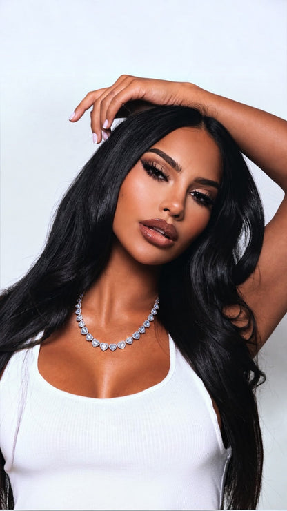A woman with long, wavy black hair models a silver rhinestone necklace and a white ribbed tank top against a light gray studio background.
